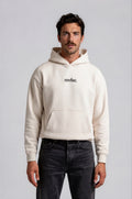 Man wearing a beige oversized hoodie with 'mvbe' logo against a plain background. ribbed sleeves and hem. warm oversized hoodie, these are uni-sex unisex products.